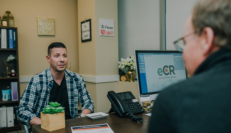 two men speaking with each other one behind a desk.